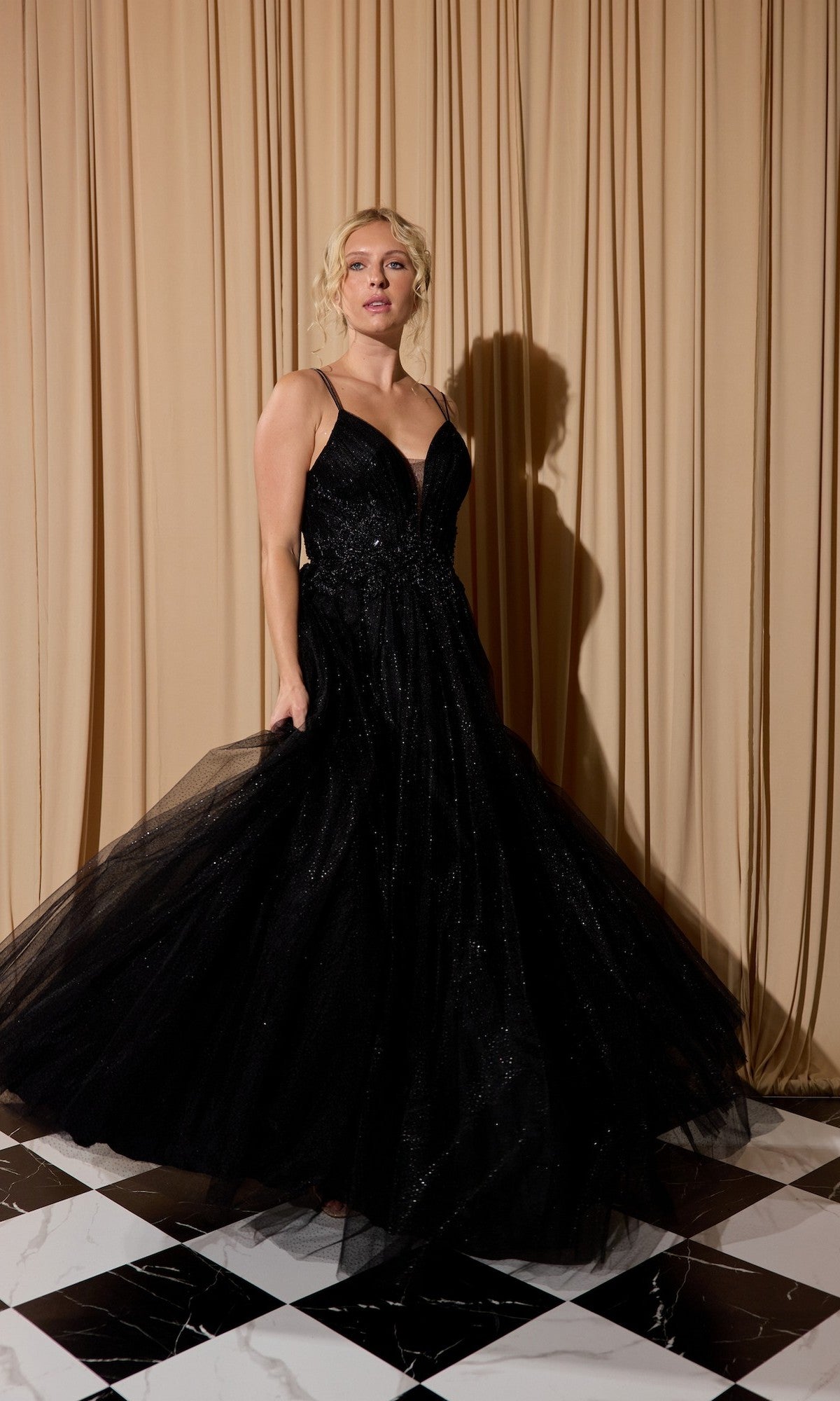 A woman wearing the Lace-Up Long Shimmer Prom Dress: Eureka 9797 in black stands on a black and white tiled floor before beige curtains.